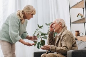 senior woman giving pills and glass of water to old man with walking stick