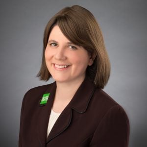 Professional headshot of Dr. Rebecca Gilbert, smiling towards the camera with shoulder length brown hair, wearing a white blouse and brown coat, with a green APDA button on her right lapel.