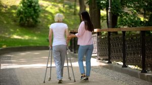 Volunteer walking with elderly woman using walker in hospital park, disability