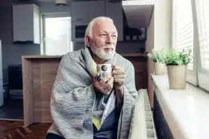 Senior men covered with blanket sitting beside heater in living room, drinking hot tea from big cup and trying to warm 