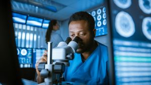 Medical Research Scientist Looking under the Microscope in the Laboratory. Neurologist Solving Puzzles of the Mind and Brain. In the Laboratory with Multiple Screens Showing MRI / CT Brain Scan Images.