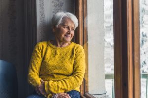Sad Caucasian Grey Haired Senior Pensioner Looking through the window with winter scenery - Smiling but Sad eyes